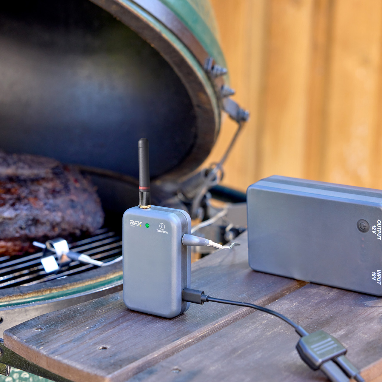 A wireless meat thermometer setup next to a grill, with a probe inserted into meat and a display unit connected to a power source.